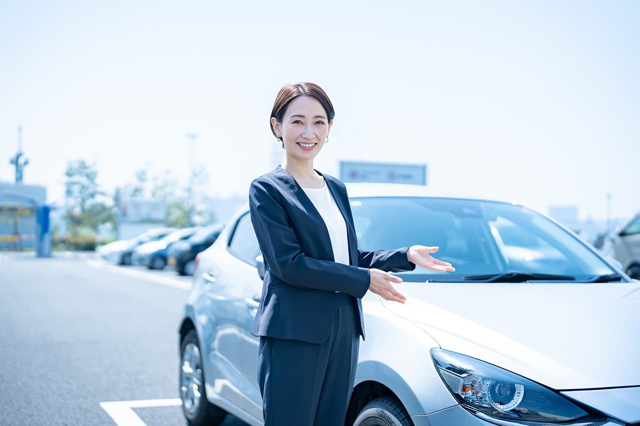 車の横に立ち、笑顔で案内するスーツ姿の女性